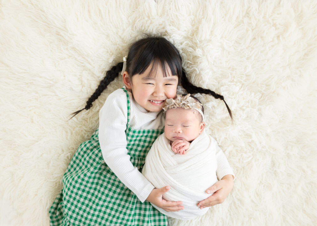 ニューボーンフォト撮影における姉妹写真。4歳のお姉ちゃんがにっこり微笑みながら、おくるみで包まれた新生児を抱っこしている様子