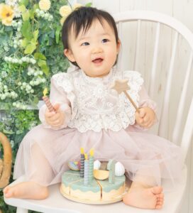 1歳の女の子がくすみピンク色のワンピースを着て、ケーキのおもちゃを前に喜んでいる様子をスタジオ撮影してる写真
