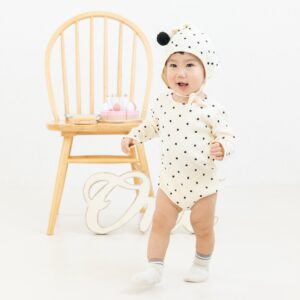 白地に黒の水玉模様のロンパースとお揃いのボンネットを着た1歳前後の男の子が、スタジオ内を楽しそうに歩いている衣装紹介写真