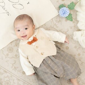 生後100日の男の子の赤ちゃんがオレンジ色の蝶ネクタイのついたロンパースを着て、仰向けに寝転んで笑顔になっている衣装の紹介写真