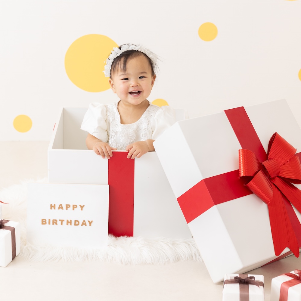 プレゼントの形をした箱から、1歳の女の子が笑顔でこちらをみている様子