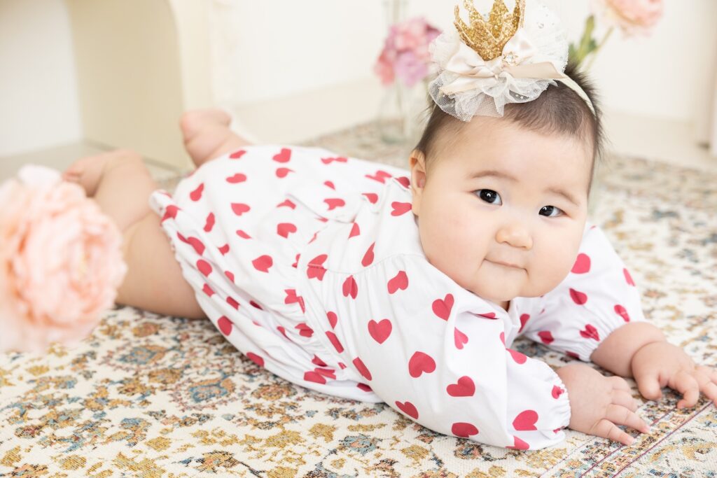 生後6ヶ月の女の子の赤ちゃんが、白地にピンクのハート模様のロンパースを着てうつ伏せポーズをしている、スタジオでのハーフバースデーの様子